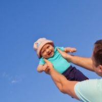 cute baby girl with hat being held up by her dad in a bright blue sky