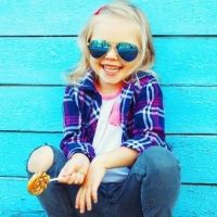 trendy little girl wearing sunglasses crouching in front of a bright blue wood wall